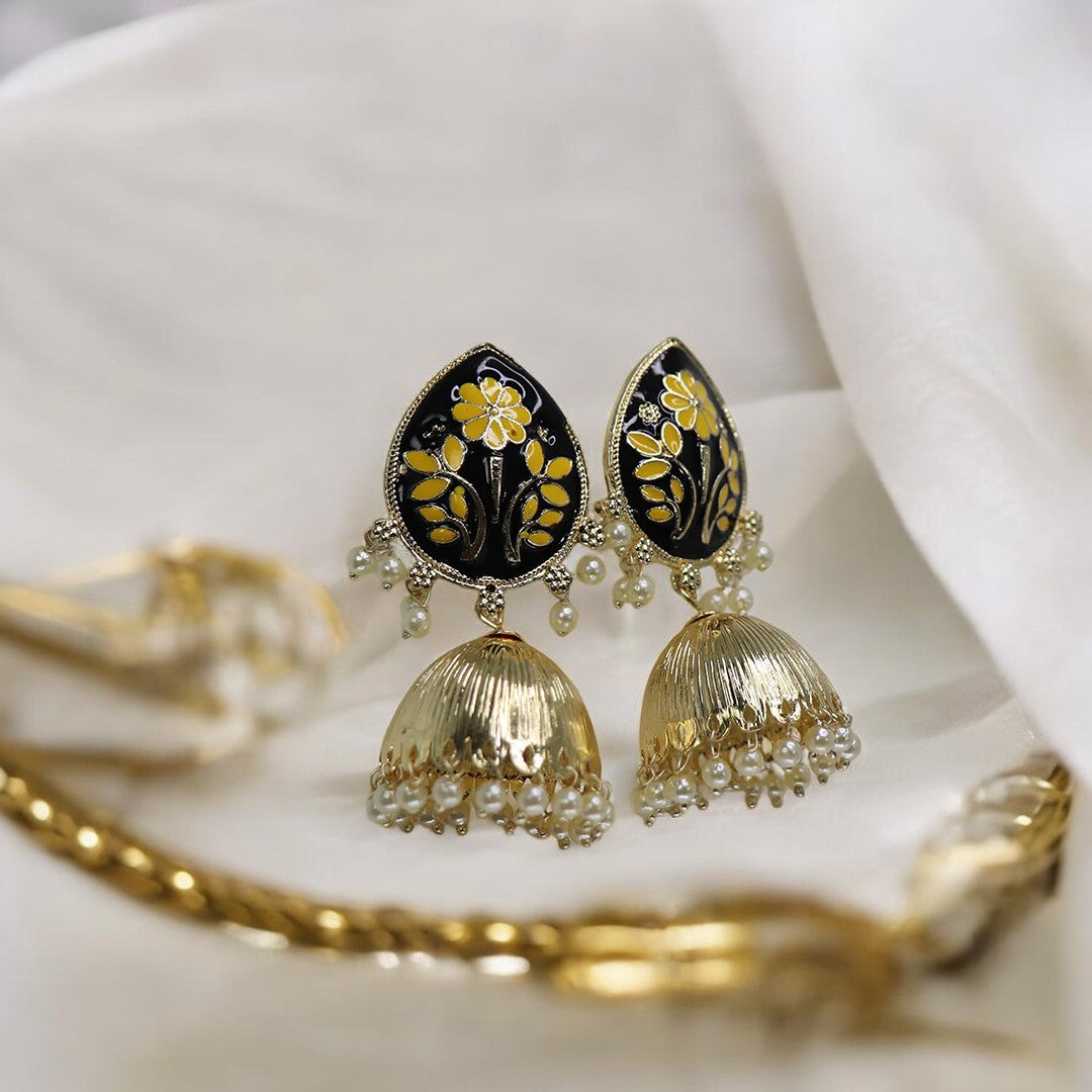A pair of black and gold colored jhumka earrings with floral patterns and dangling embellishments, displayed on a white fabric surface.