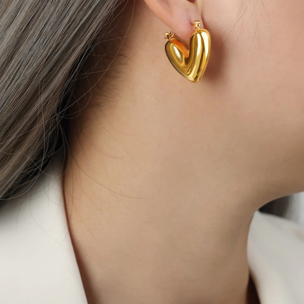 A close-up image of a person wearing 18K gold plated heart-shaped hoop earrings.