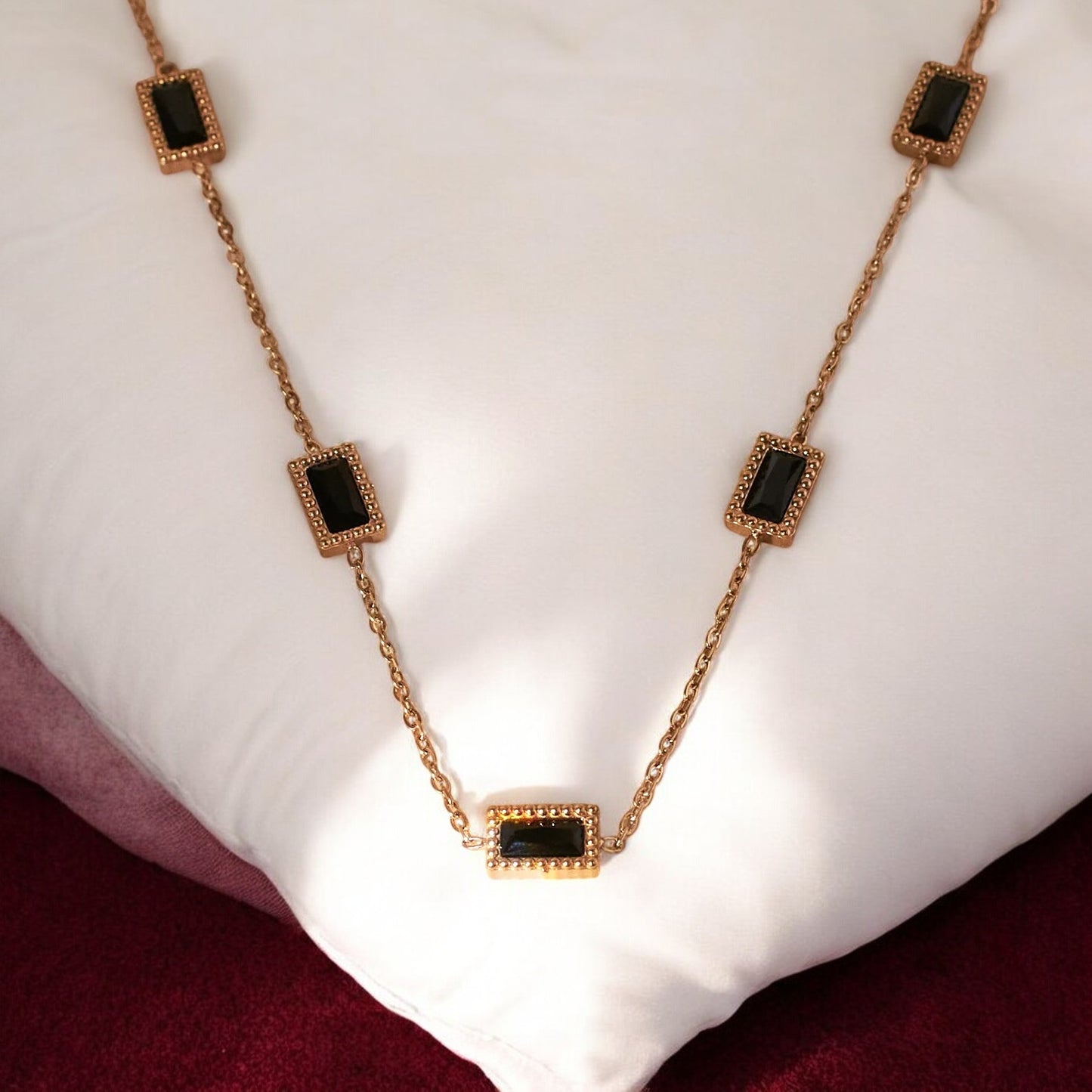 A gold-colored pendant necklace with black square accents, displayed on a white cushion.