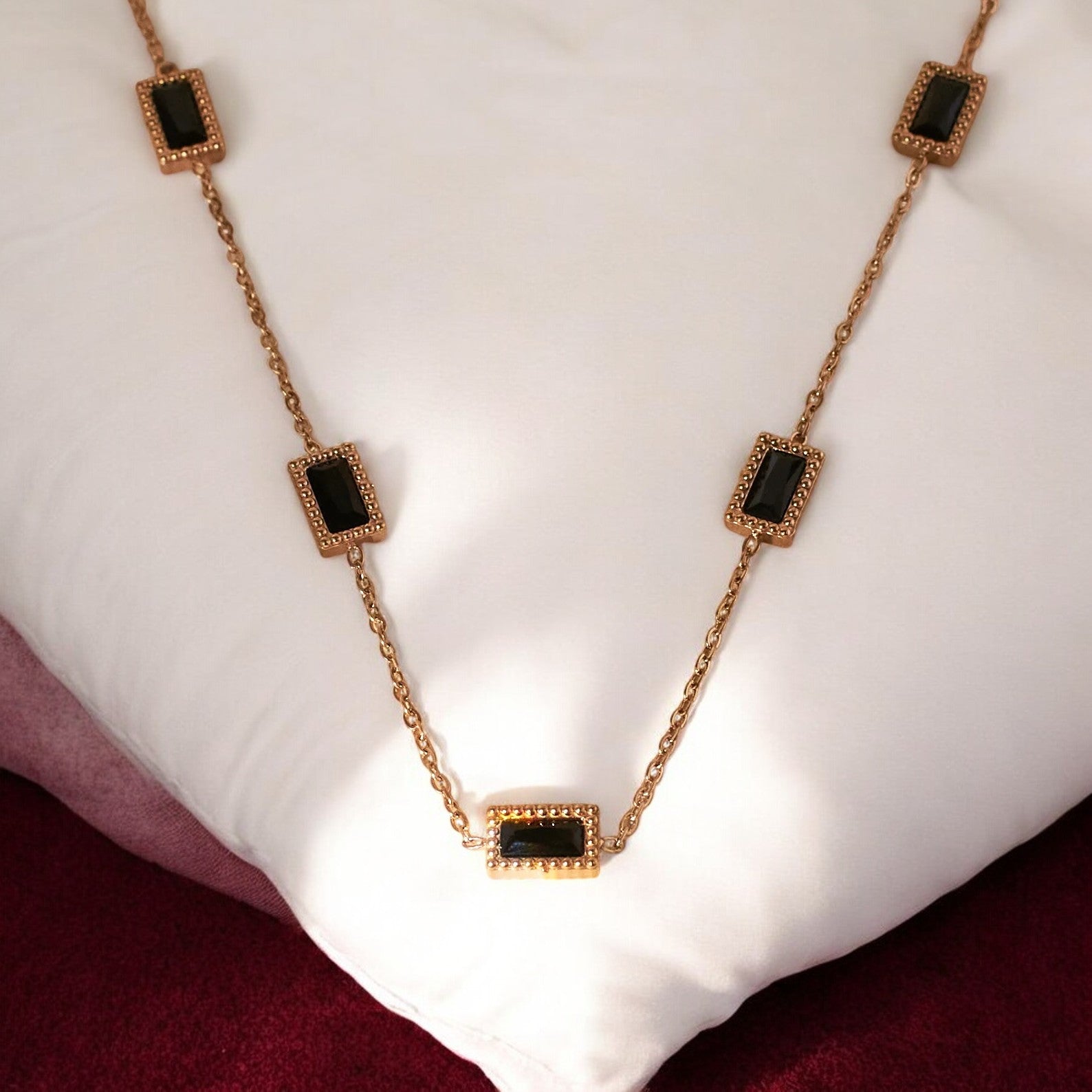 A gold-colored pendant necklace with black square accents, displayed on a white cushion.