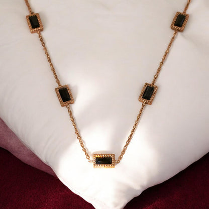 A gold-colored pendant necklace with black square accents, displayed on a white cushion.