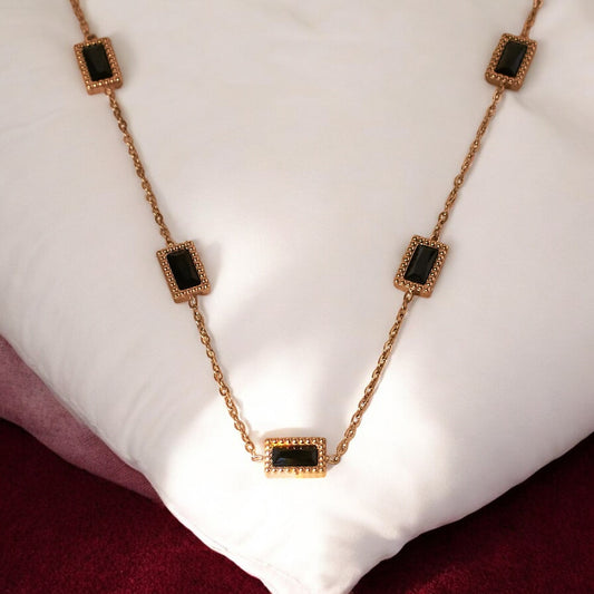 A gold-colored pendant necklace with black square accents, displayed on a white cushion.