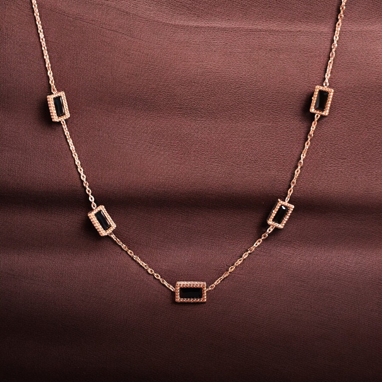 A rose gold chain necklace with black stone accents displayed against a brown background.