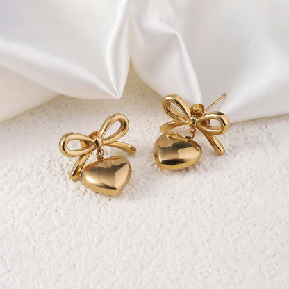 This image displays a pair of elegant earrings, crafted in 18kt gold. Each earring features a charming design consisting of a heart-shaped bell dangled from a beautifully tied bow.