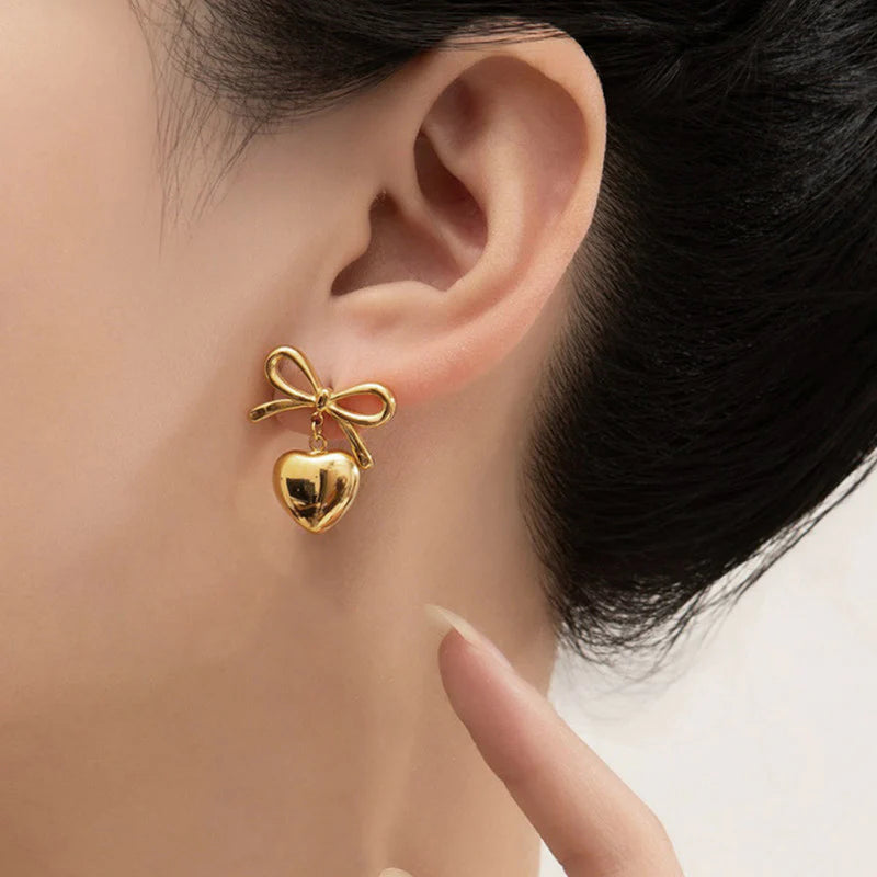 A close-up image of a person wearing 18Kt gold-plated earrings with a heart and bow design.
