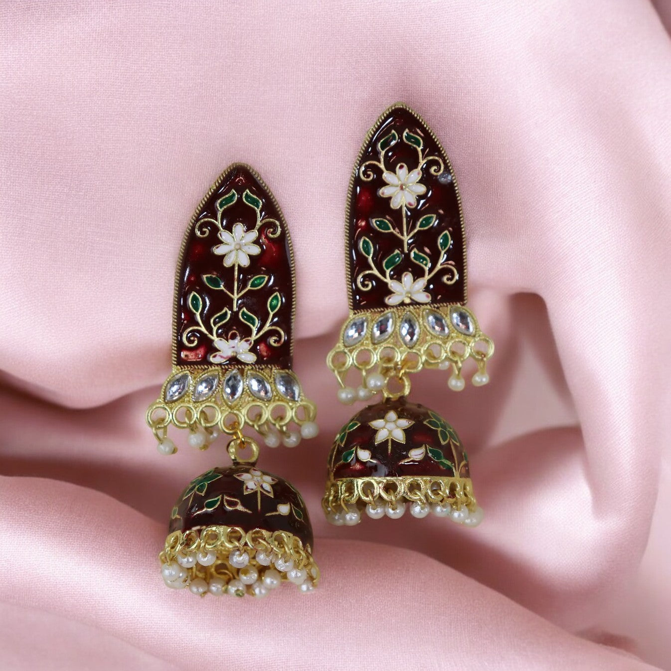 A pair of maroon meenakari earrings with gold accents and white pearls, displayed on a pink fabric surface.