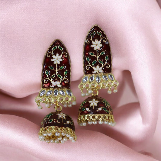 A pair of maroon meenakari earrings with gold accents and white pearls, displayed on a pink fabric surface.