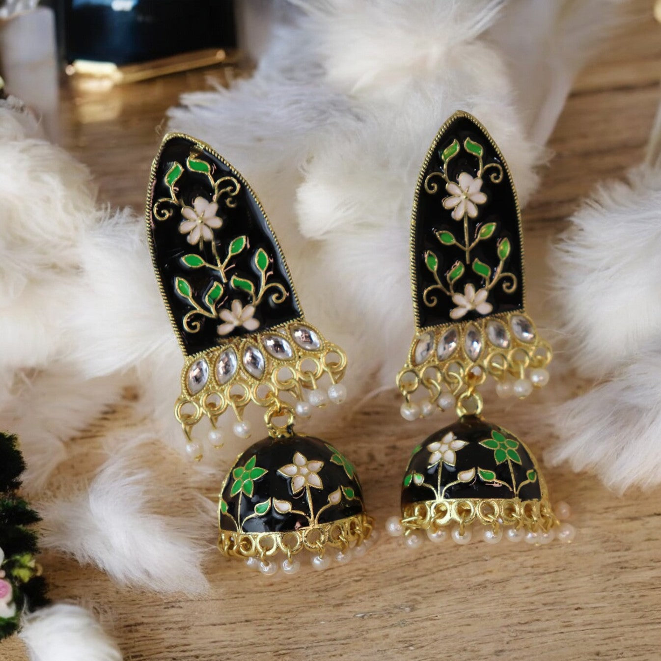 A pair of black and green meenakari earrings with gold accents and pearl embellishments, displayed on a flat surface with white furry objects around.