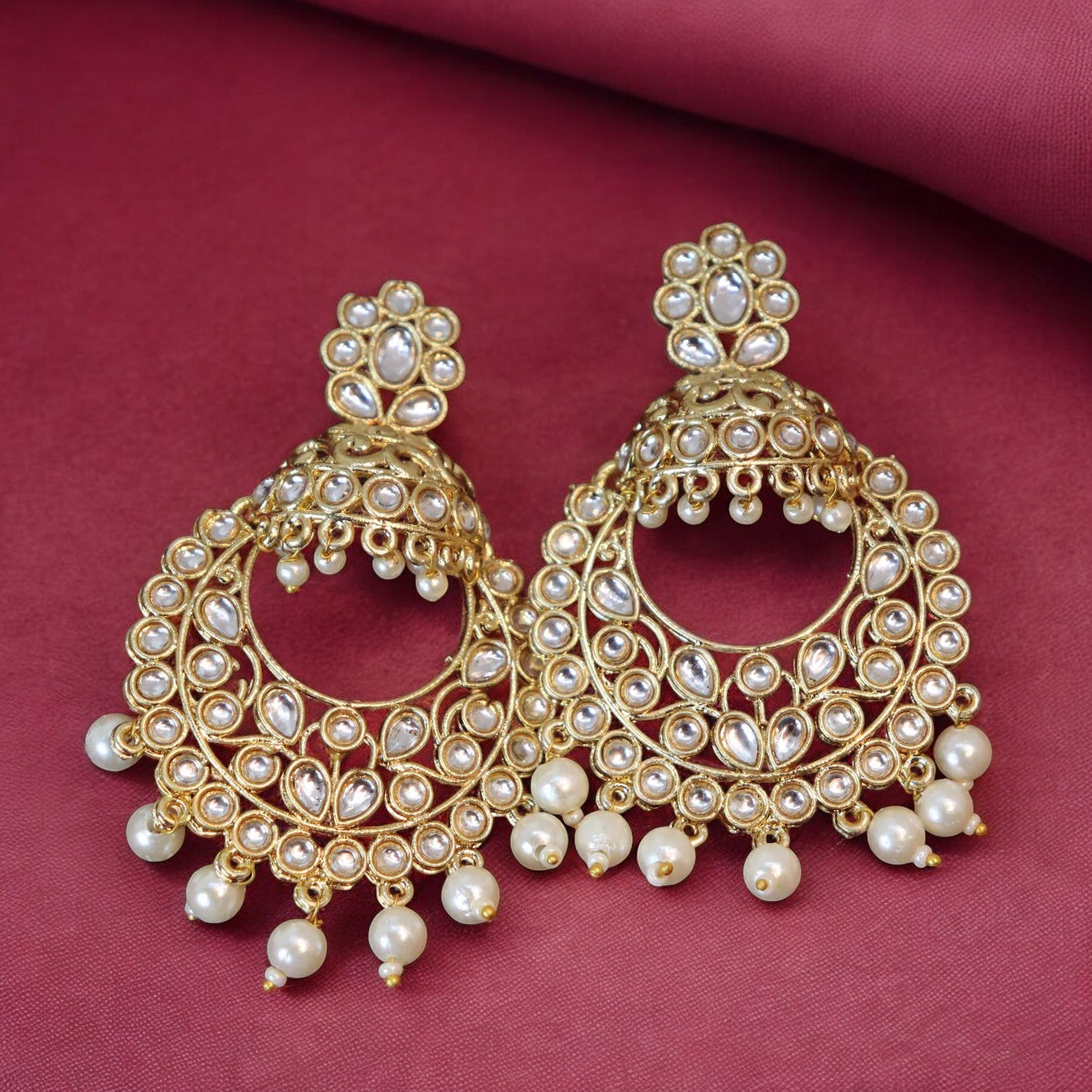 A pair of golden kundan chandbali earrings with pearl embellishments on a pink background.