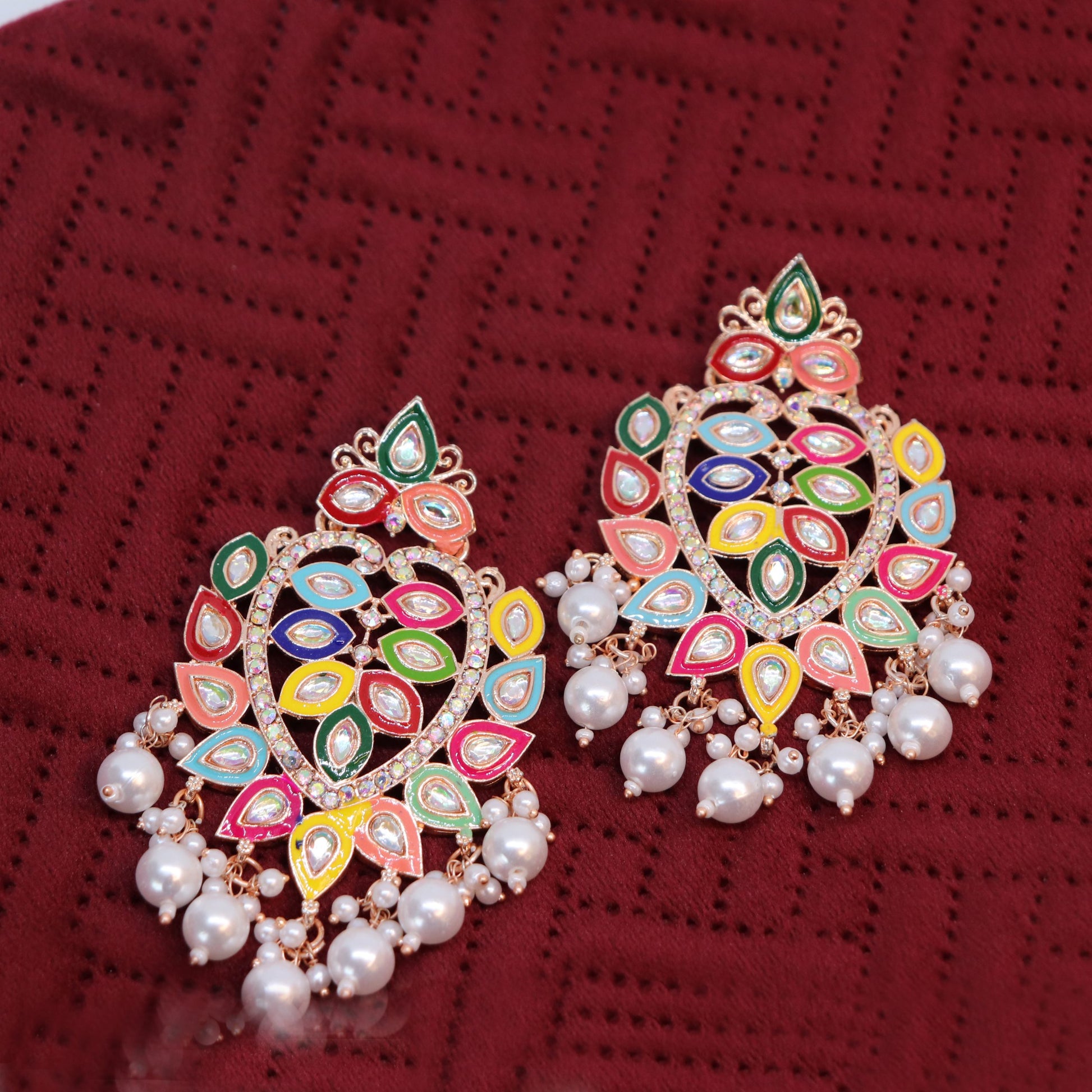 A pair of multicolored Chandbali earrings with pearl drops, displayed against a red background.
