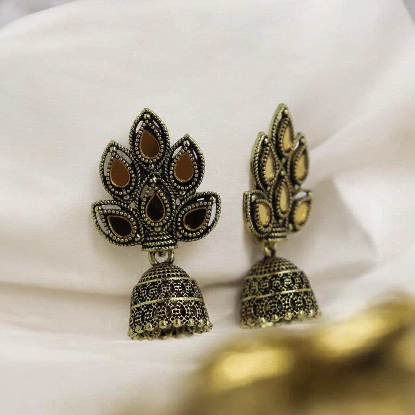 A pair of golden jhumki earrings with intricate leaf patterns, displayed on a white fabric surface.