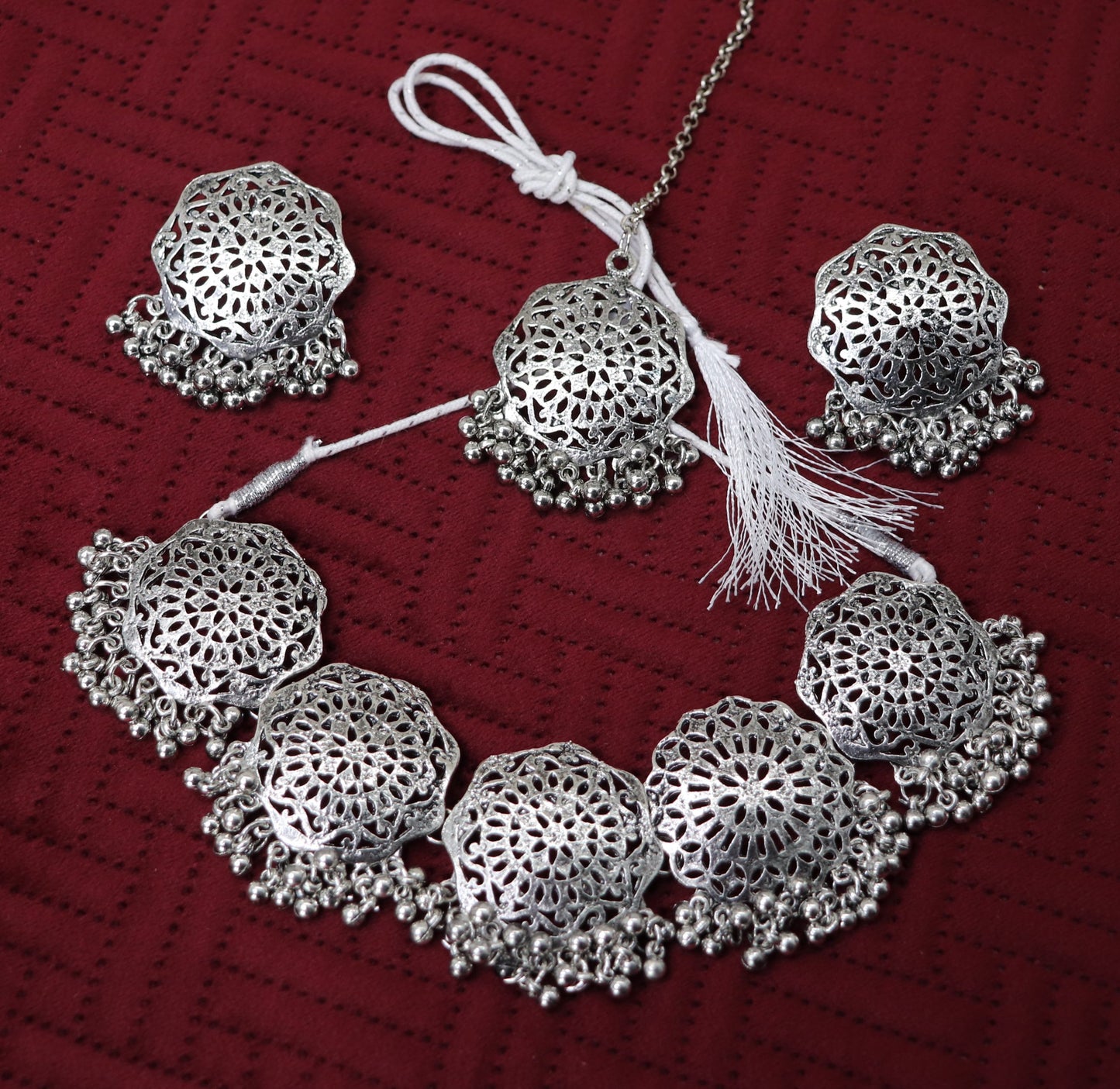 A silver Boho choker necklace set with intricate geometric patterns and small dangling beads, displayed on a red background.