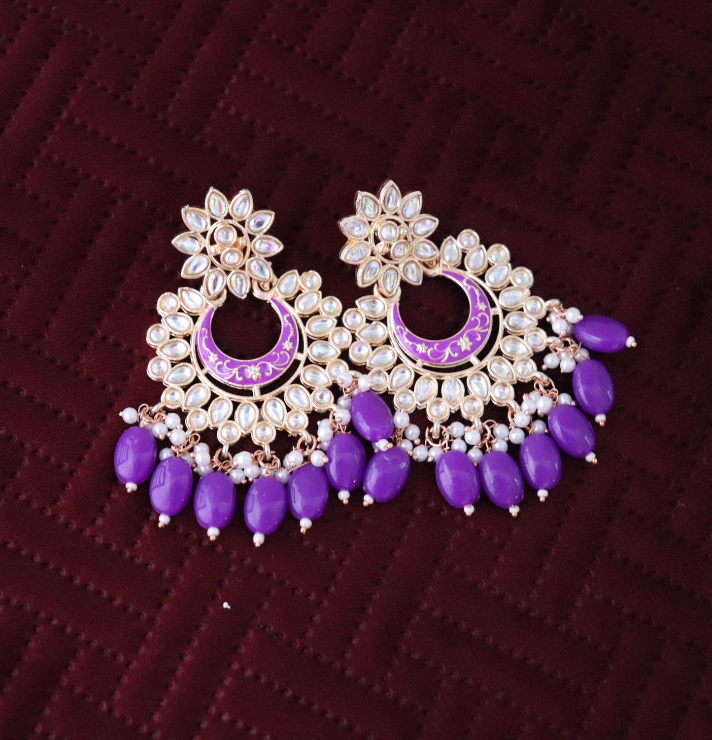 A pair of gold-colored metal Chandbali earrings featuring purple beads and intricate designs, displayed against a maroon background.