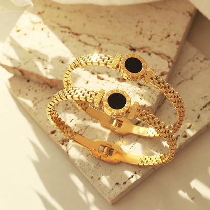 Two gold bracelets with black accents are displayed on a textured, light-colored surface, catching the light to highlight their shine and design.