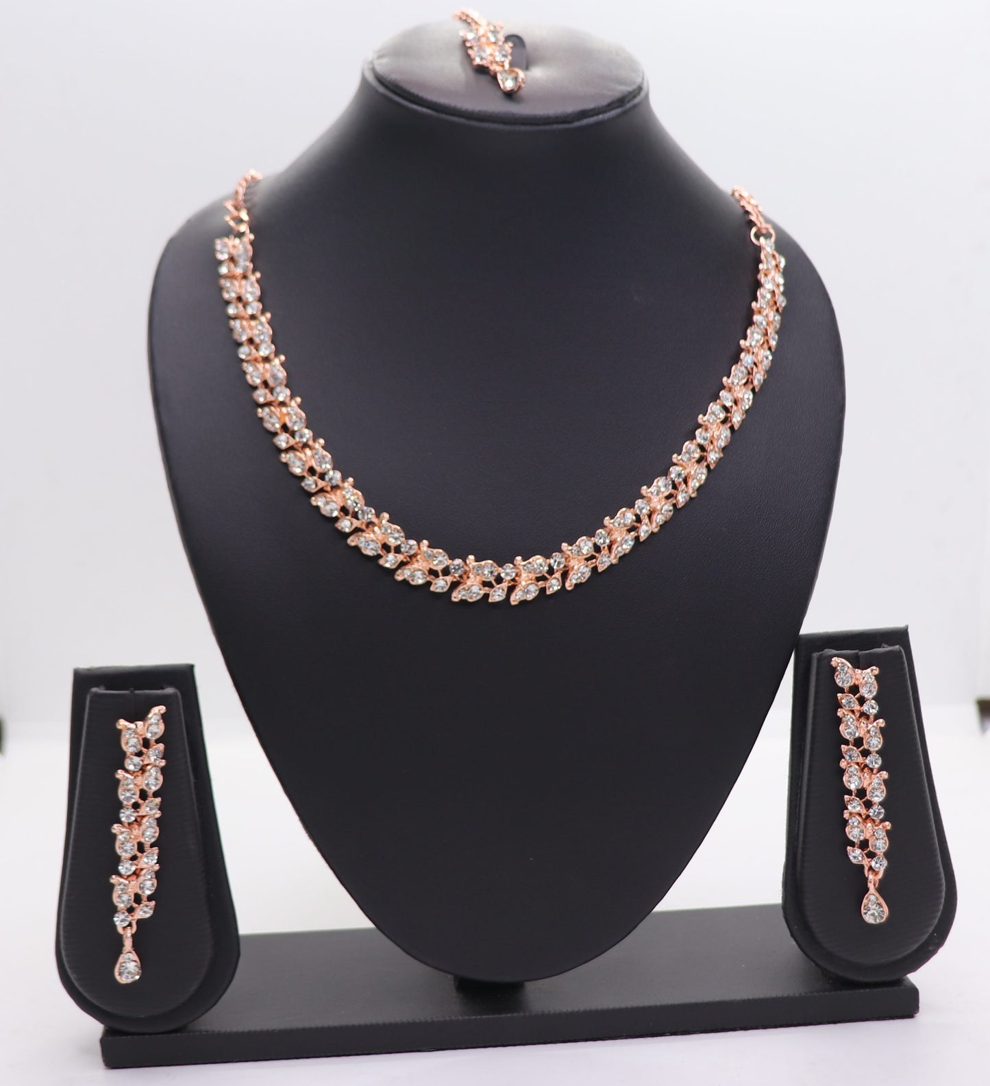 A set of AD chokers, including a necklace, earrings, and a mangtikka, displayed on a black mannequin. The jewelry features a rose gold color with floral and leaf patterns adorned with rhinestones.