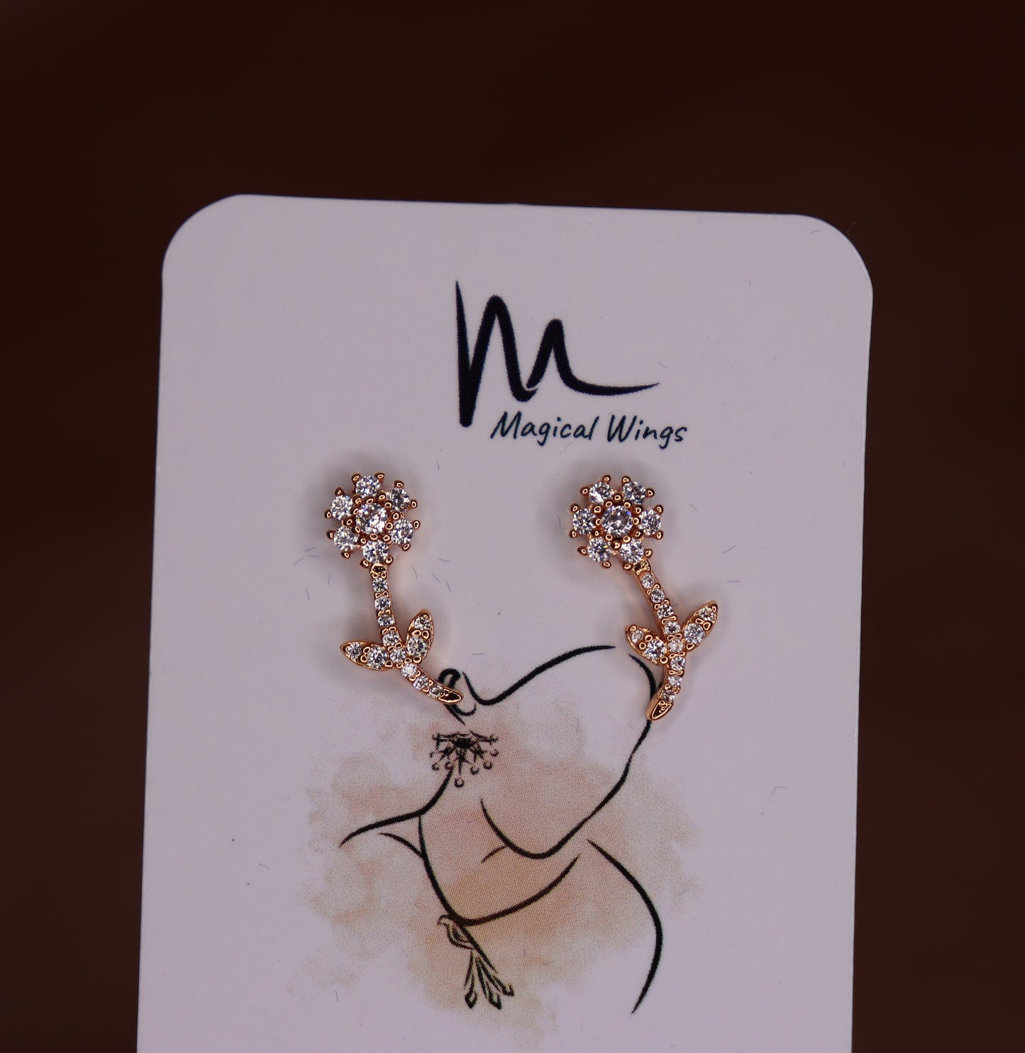 A pair of stud earrings with a floral pattern in a silver tone, displayed on a branded card with 'Magical Wings' logo.