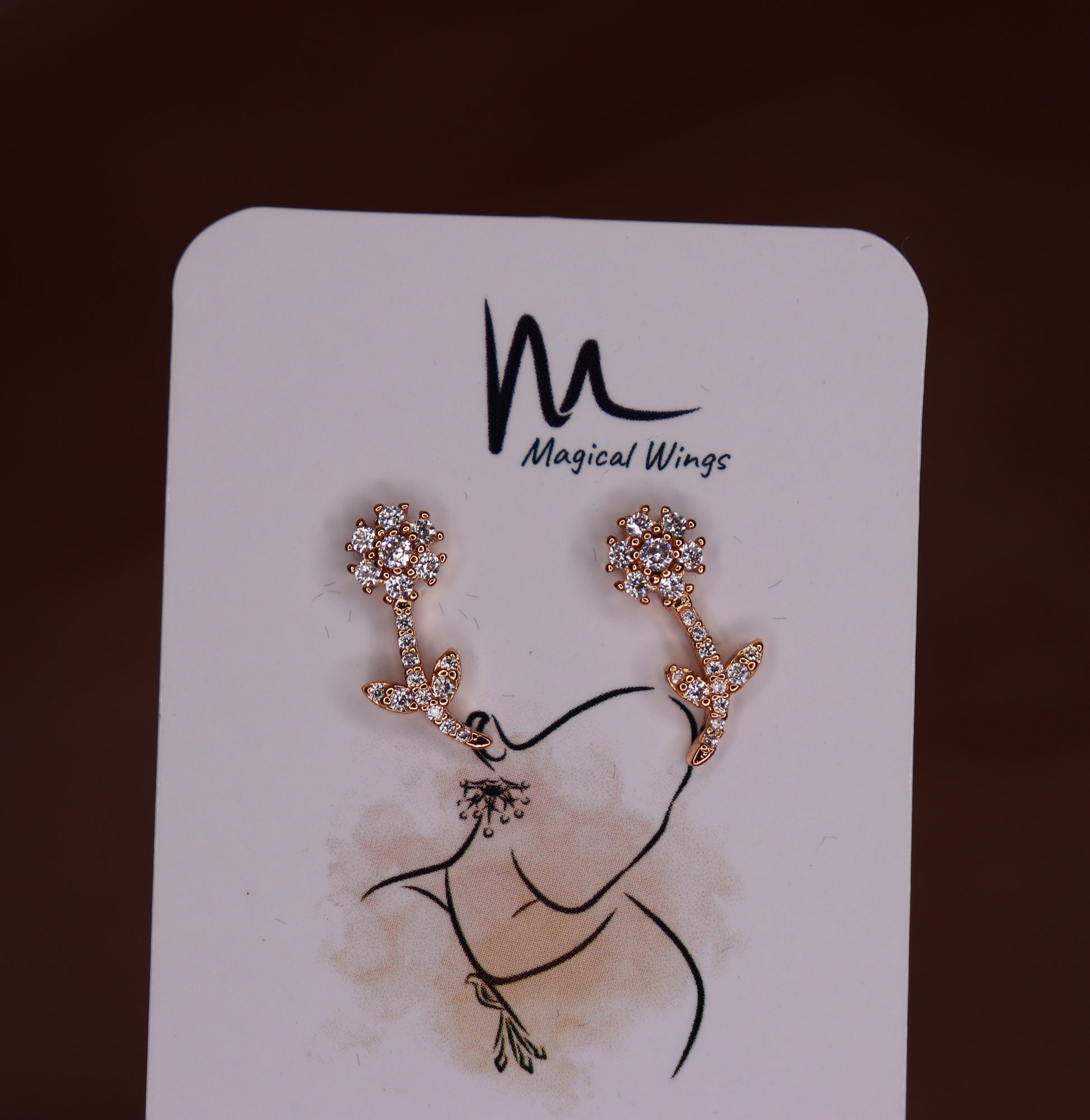 A pair of stud earrings with a floral pattern in a silver tone, displayed on a branded card with 'Magical Wings' logo.