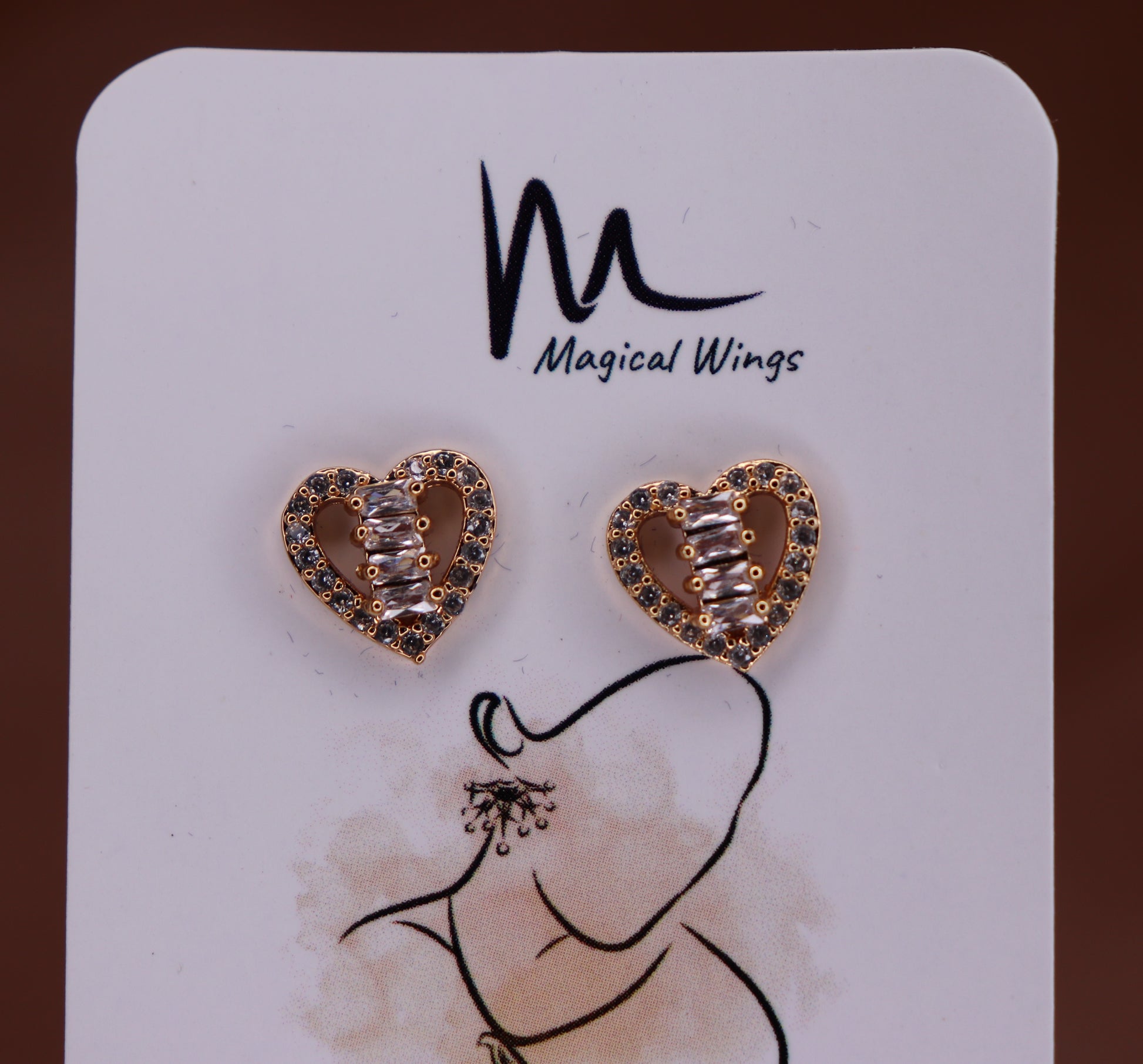 A pair of heart-shaped stud earrings with AD embellishments, displayed on a card with the brand name Magical Wings.