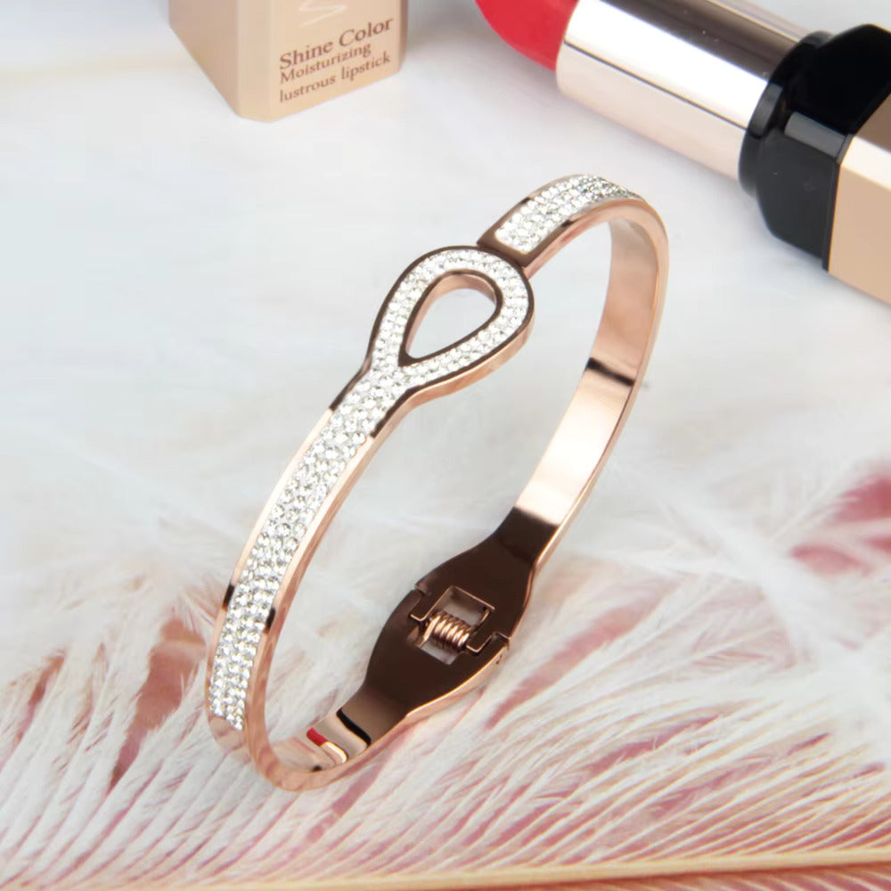 A rose gold bangle bracelet with link design and encrusted with zirconia stones, displayed alongside cosmetic products.