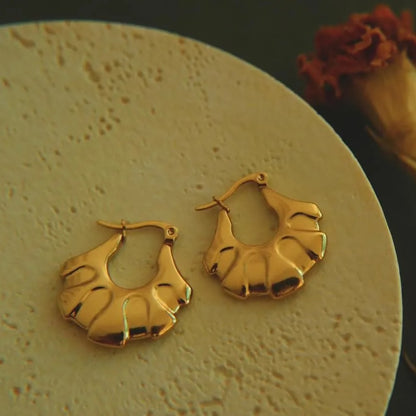 A pair of 18kt gold hoop earrings displayed on a beige surface, with a textured design.