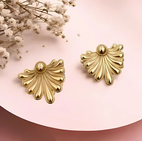 A pair of gold stud earrings with a textured, shell-like design, displayed on a pink background with some white flowers.