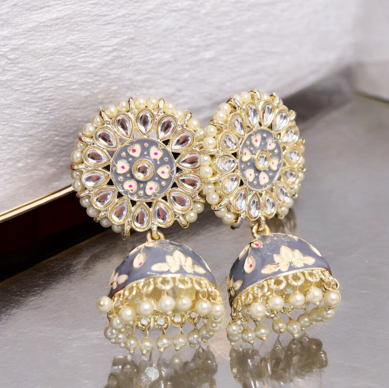 A pair of gold-colored jhumka earrings with grey and multicolored stone embellishments, displayed against a reflective background.