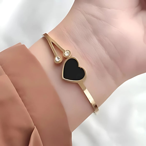 A gold bangle bracelet with a black heart-shaped stone and small diamond-like stones on the side, displayed on a person's wrist.