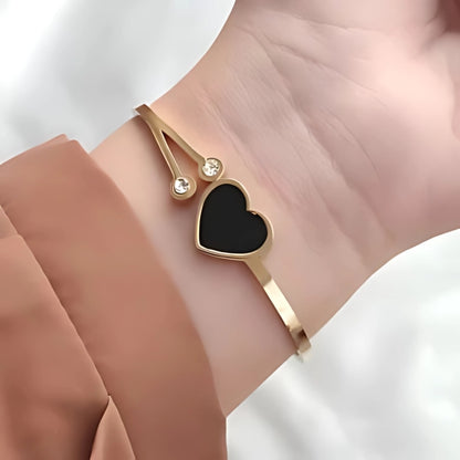 A gold bangle bracelet with a black heart-shaped stone and small diamond-like stones on the side, displayed on a person's wrist.