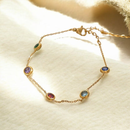 A 18K thin chain gold bracelet with multiple colored charms, displayed on a beige fabric surface.