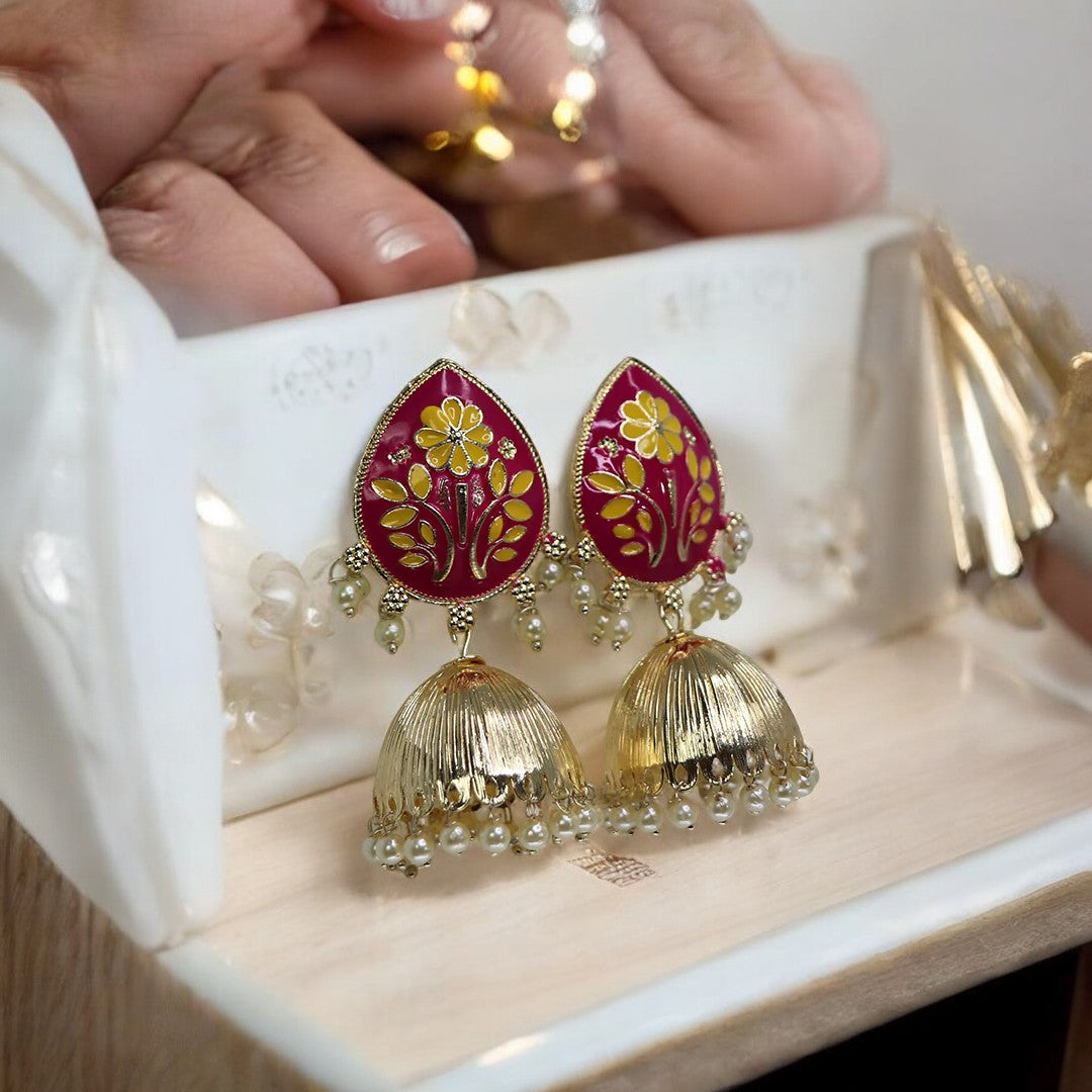 A pair of magenta and gold floral patterned jhumka earrings displayed in a white box with intricate gold detailing.