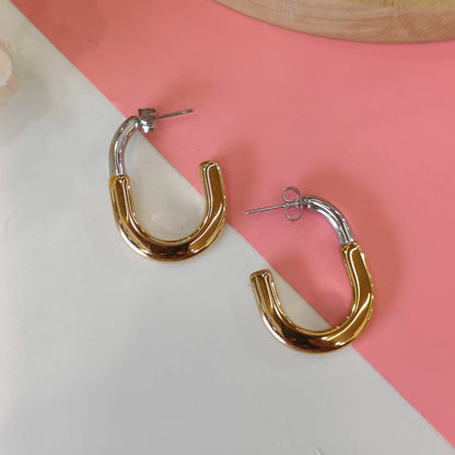 This image features a pair of elegant 18kt gold hoop earrings displayed against a two-tone background split down the middle, with the upper half in pink and the lower half in a neutral shade. The earrings exhibit a modern and sleek design, with a metallic finish that enhances their sophisticated appeal.