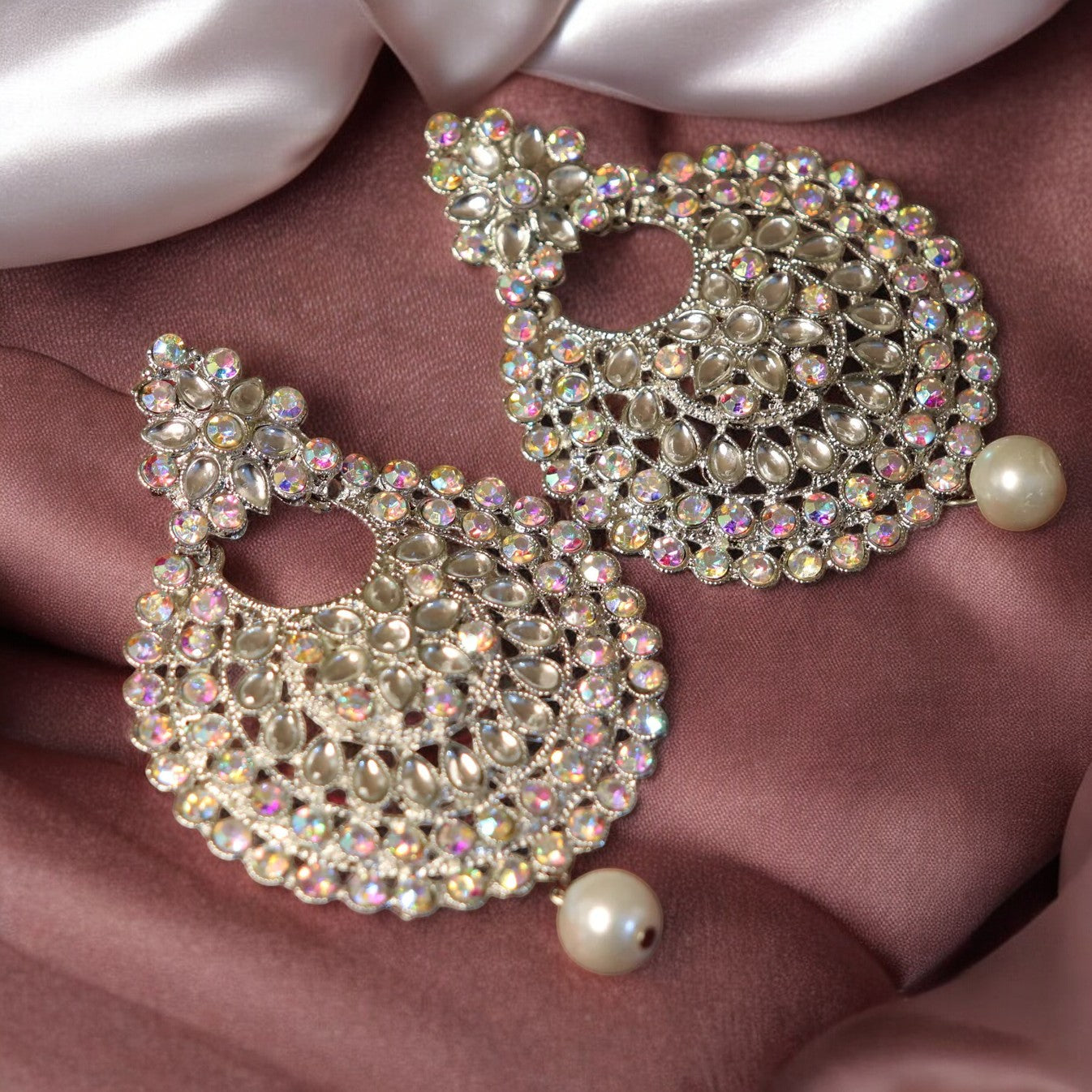 A pair of silver Chandbali earrings adorned with multicolored crystals and pearls, displayed on a purple fabric.